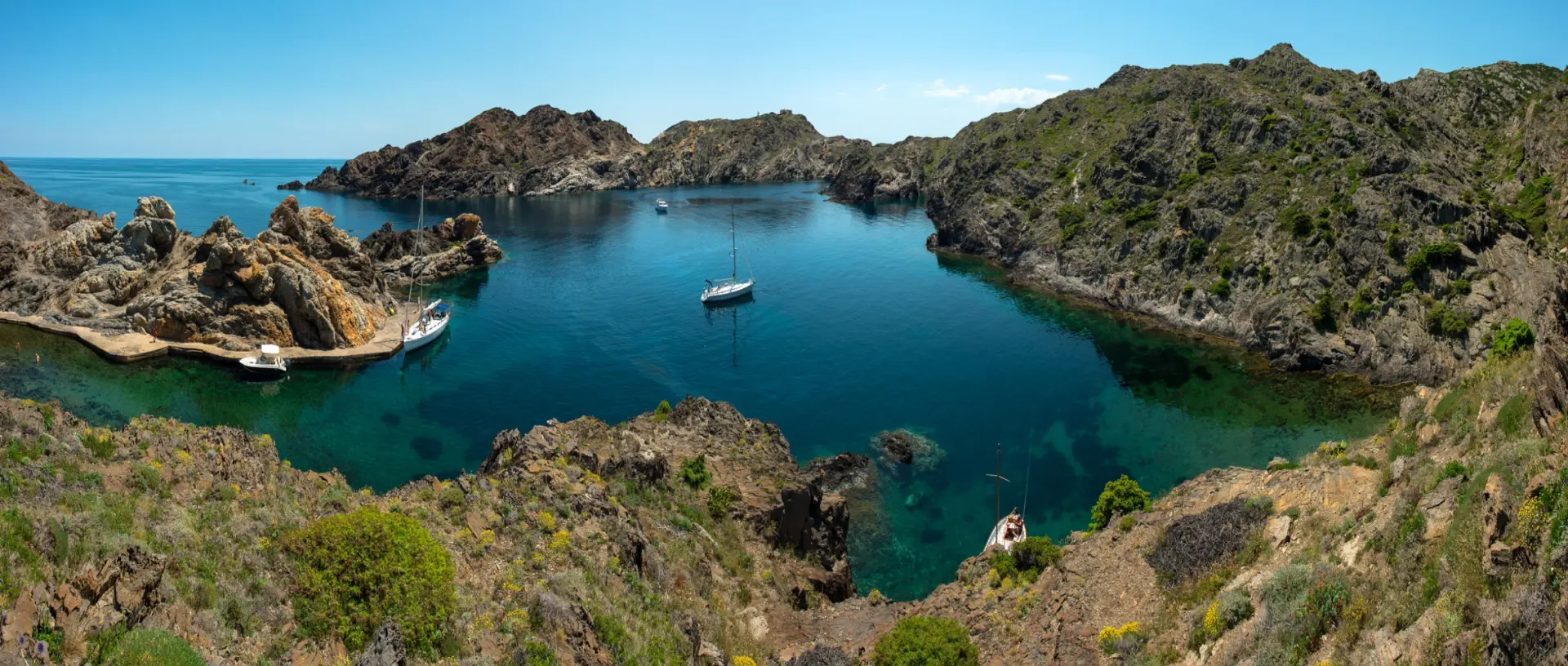 Cala d’aigua turquesa al Cap de Creus, amb velers ancorats entre roques daurades